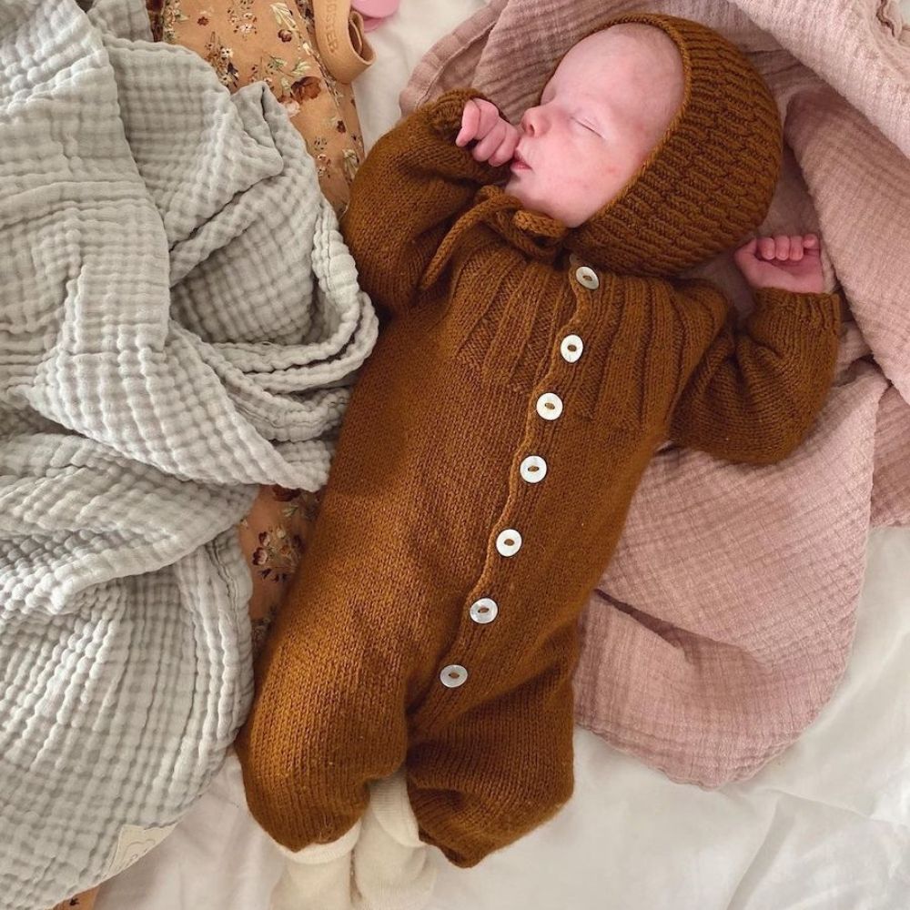 A baby dressed in a brown knitted outfit lies on a bed wearing the anker bonnet, exuding comfort and tranquility in a serene setting.