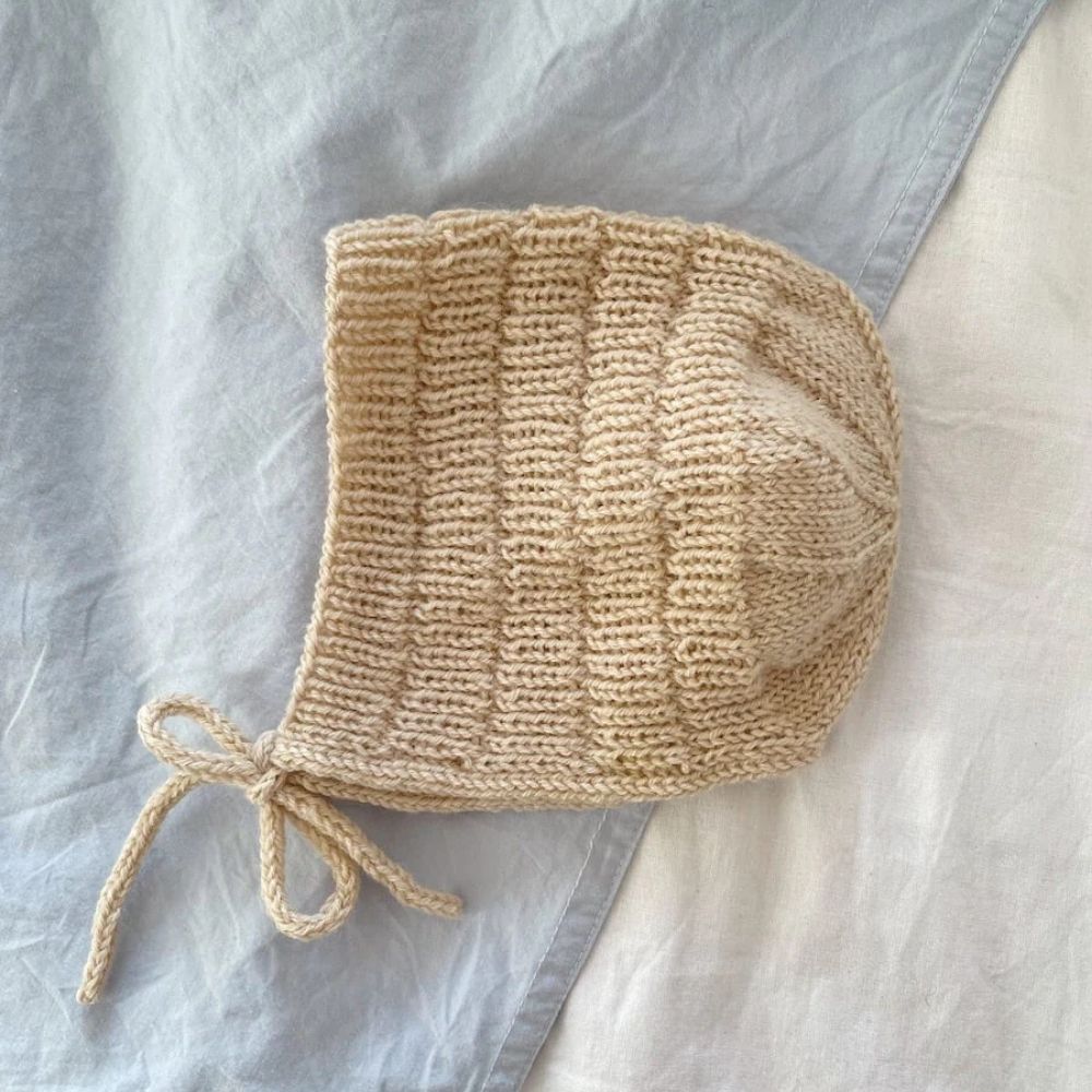 A knitted bonnet displayed on a bed, highlighting its ribbed design and finished with an i-cord edge and ties.