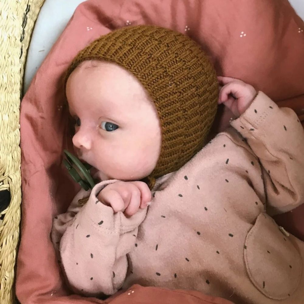 A baby wearing a knitted hat and a pink shirt, featuring a delightful design with tie-strings for a secure fit.