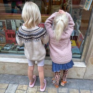 Two small girls look intently at books in a window, showcasing their interest in reading and exploration, wearing their warm sweaters.