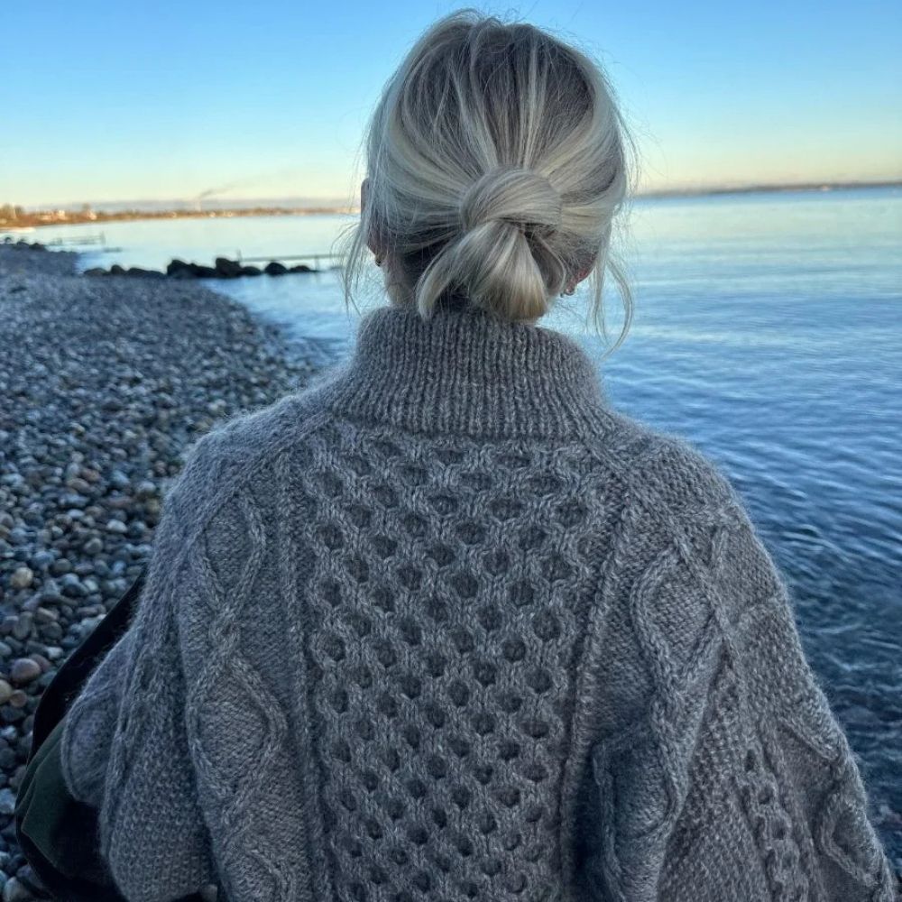 A woman wearing a cozy sweater stands on the beach, highlighting the detailed design of the Dagmar Zipper Sweater.