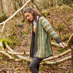 A woman in a knitted cardigan called Airadale by Rowan stands surrounded by lush greenery in a peaceful forest environment.