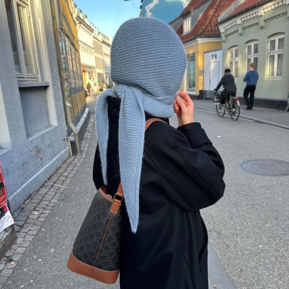 A woman in a black coat and a light blue knitted headscarf walks down a city street, holding her bag and looking thoughtfully.