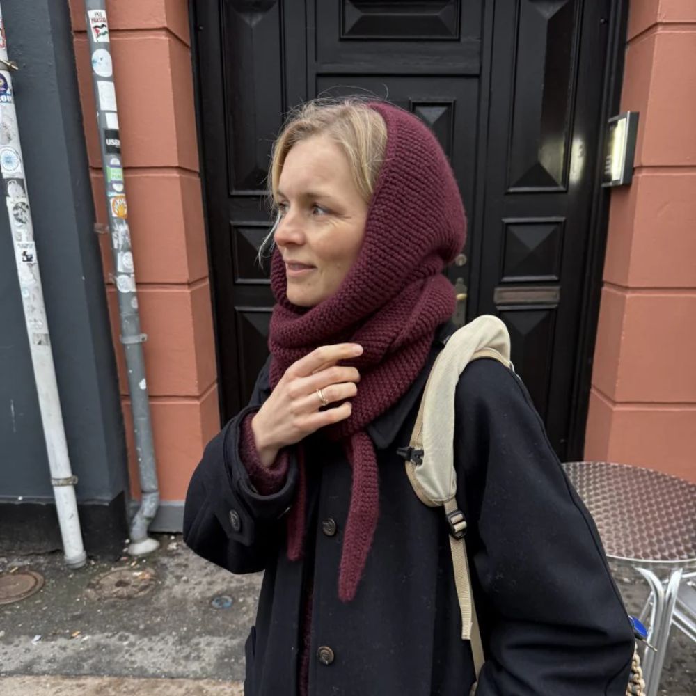 A person wearing a dark coat and a burgundy scarf stands near a black door, casually holding their hand up.
