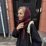 A person wearing a dark coat and a burgundy scarf stands near a black door, casually holding their hand up.