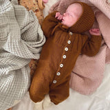 A baby dressed in a brown knitted outfit lies on a bed wearing the anker bonnet, exuding comfort and tranquility in a serene setting.