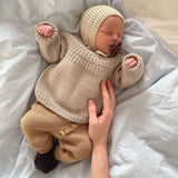A baby in a knitted sweater and anker bonnet hat peacefully rests on a bed, showcasing the intricate design of the bonnet and sweater.
