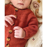 A baby in a knitted sweater lies on a bed, showcasing the intricate design of Anker’s Onesie with ribbed details.