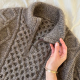 A woman holds a zipped sweater, showcasing intricate cable and texture patterns, designed for a stylish, cozy fit.