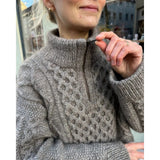 A woman models a cable knit sweater, showcasing intricate patterns and a stylish zipper detail at the front opening.
