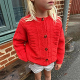A little girl in a red sweater and white shorts, smiling joyfully, showcasing a playful and vibrant outfit.