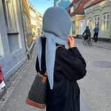 A woman in a black coat and a light blue knitted headscarf walks down a city street, holding her bag and looking thoughtfully.