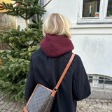 A woman with wavy blonde hair, wearing a black coat and burgundy cowl, walks past a tree and a window. A crossbody bag is slung over her shoulder.