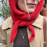 A close-up of a woman in a tan coat wearing a bright red knitted head wrap with a delicate necklace featuring a small charm.