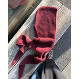 A cozy maroon knitted hat with two long ties rests on a wooden bench, with a person's feet in brown shoes beside it.